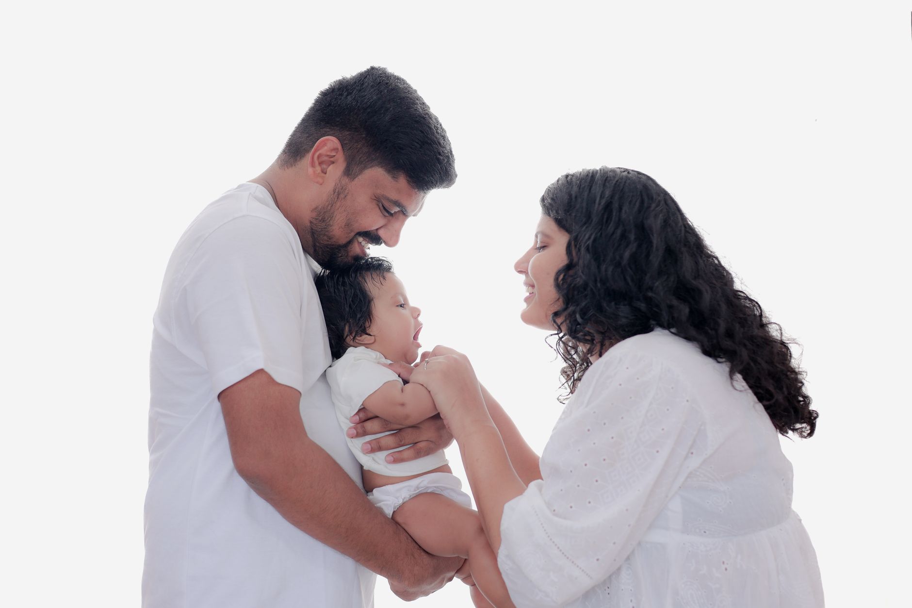 An Indian father and mother, both dressed in white, smiling and holding their newborn baby together in a bright studio setting