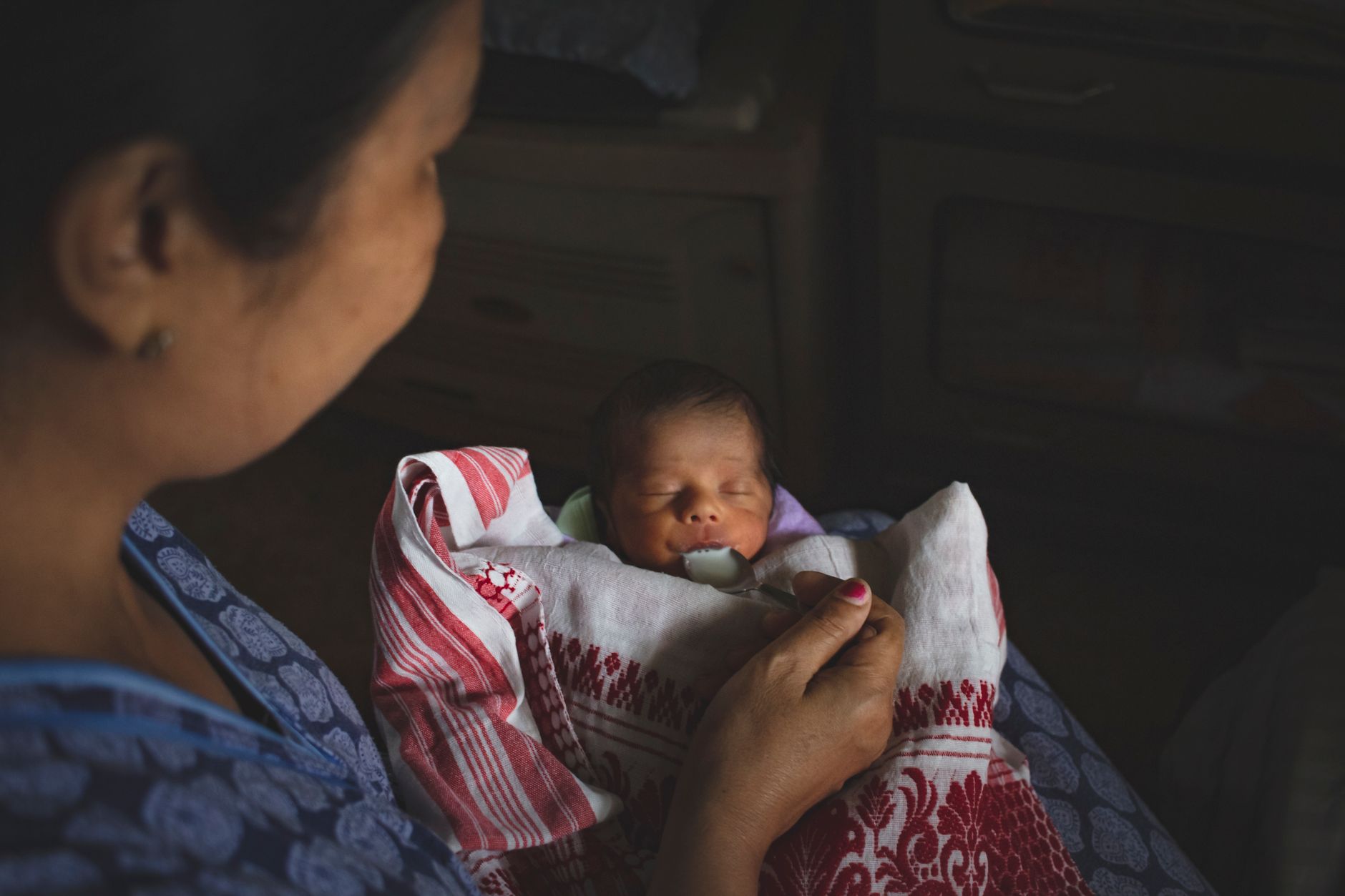 An Indian mother spoon-feeding her sleeping newborn, wrapped in a traditional gamusa cloth, in a dim, quiet room