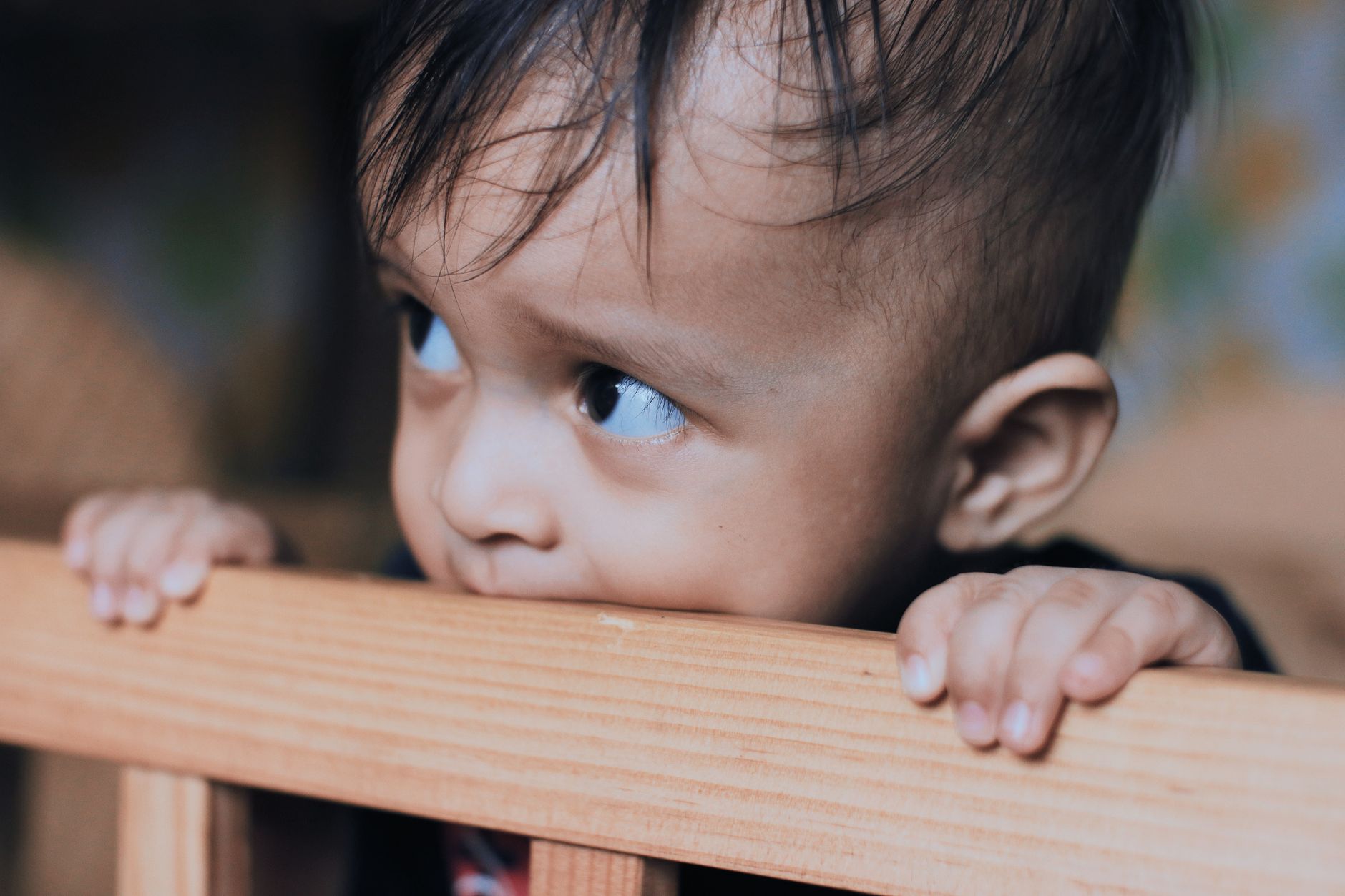 Toddler standing in a wooden crib, gripping the top rail with both hands while peeking over it