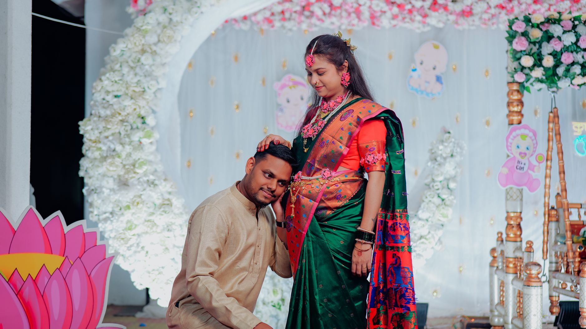 A pregnant woman in a green and orange saree and her partner, kneeling beside her, at a decorated Indian baby shower stage with floral arches and balloon decor