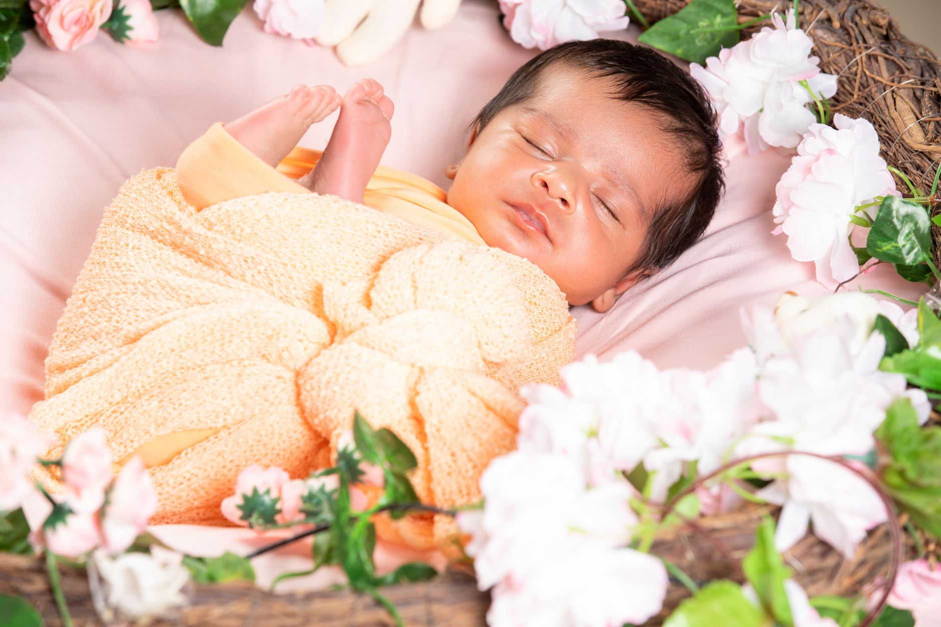 Indian newborn sleeping peacefully wrapped in an orange knit blanket inside a floral basket decorated with pink and white flowers, photographed in Bengaluru