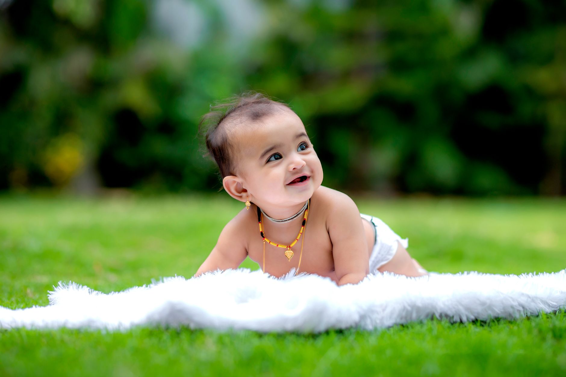 An Indian baby wearing a white diaper, smiling while lying on a blanket on grass outdoors