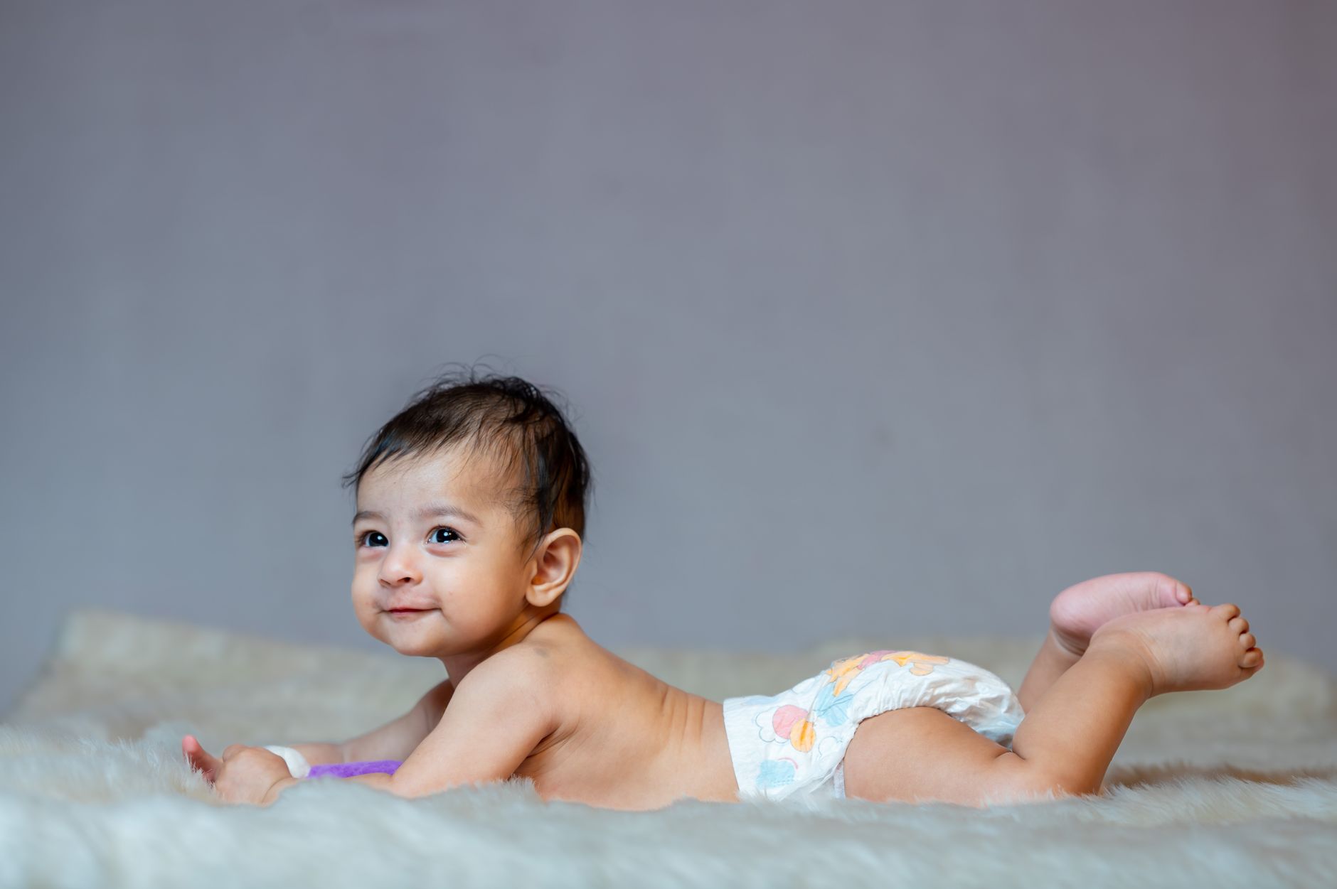 An Indian baby lying on their tummy on a soft blanket, wearing a diaper, smiling