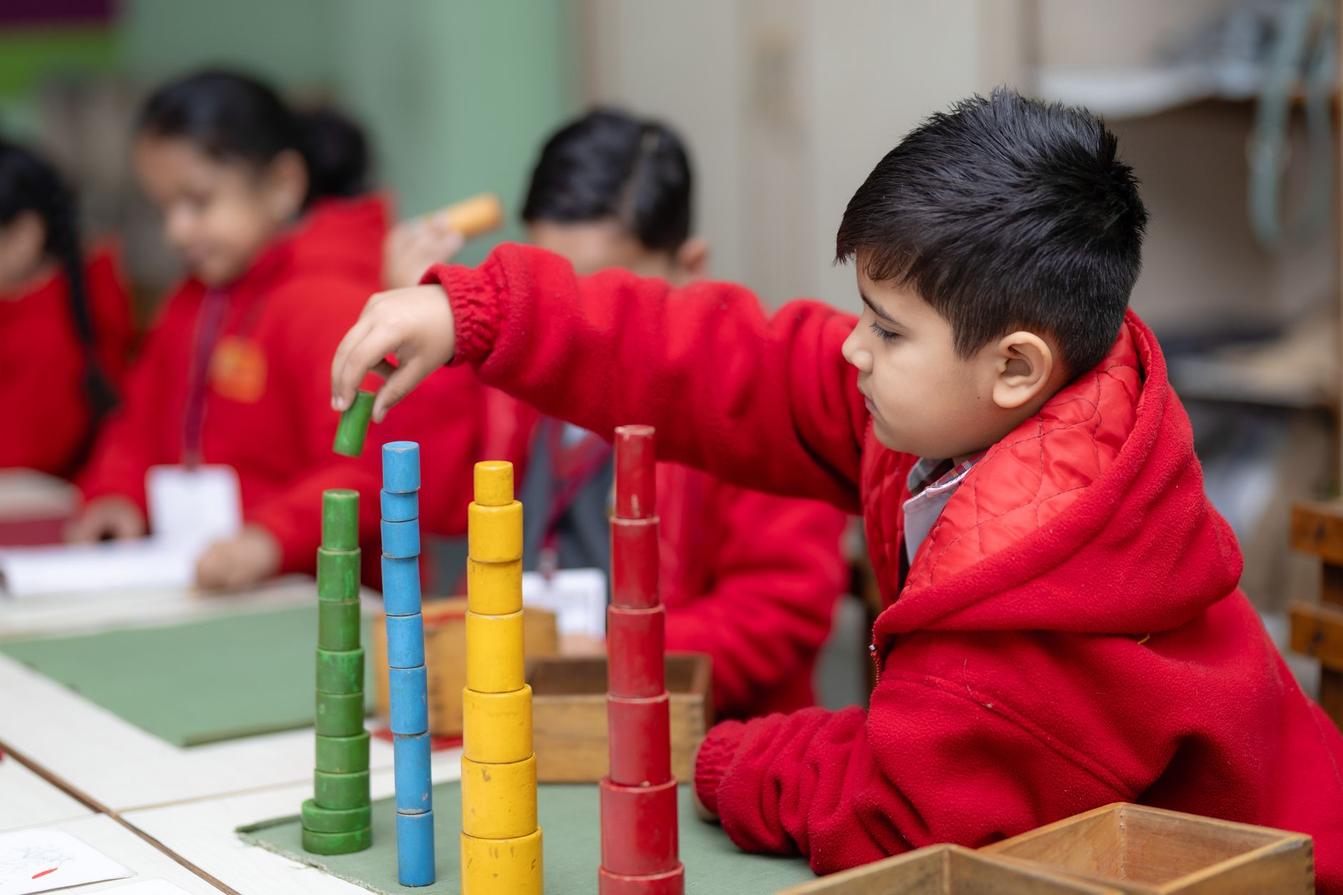 A young boy in a red uniform stacking colourful wooden blocks at a desk in a clean, well-lit Indian Montessori classroom, with classmates working at nearby desks.
