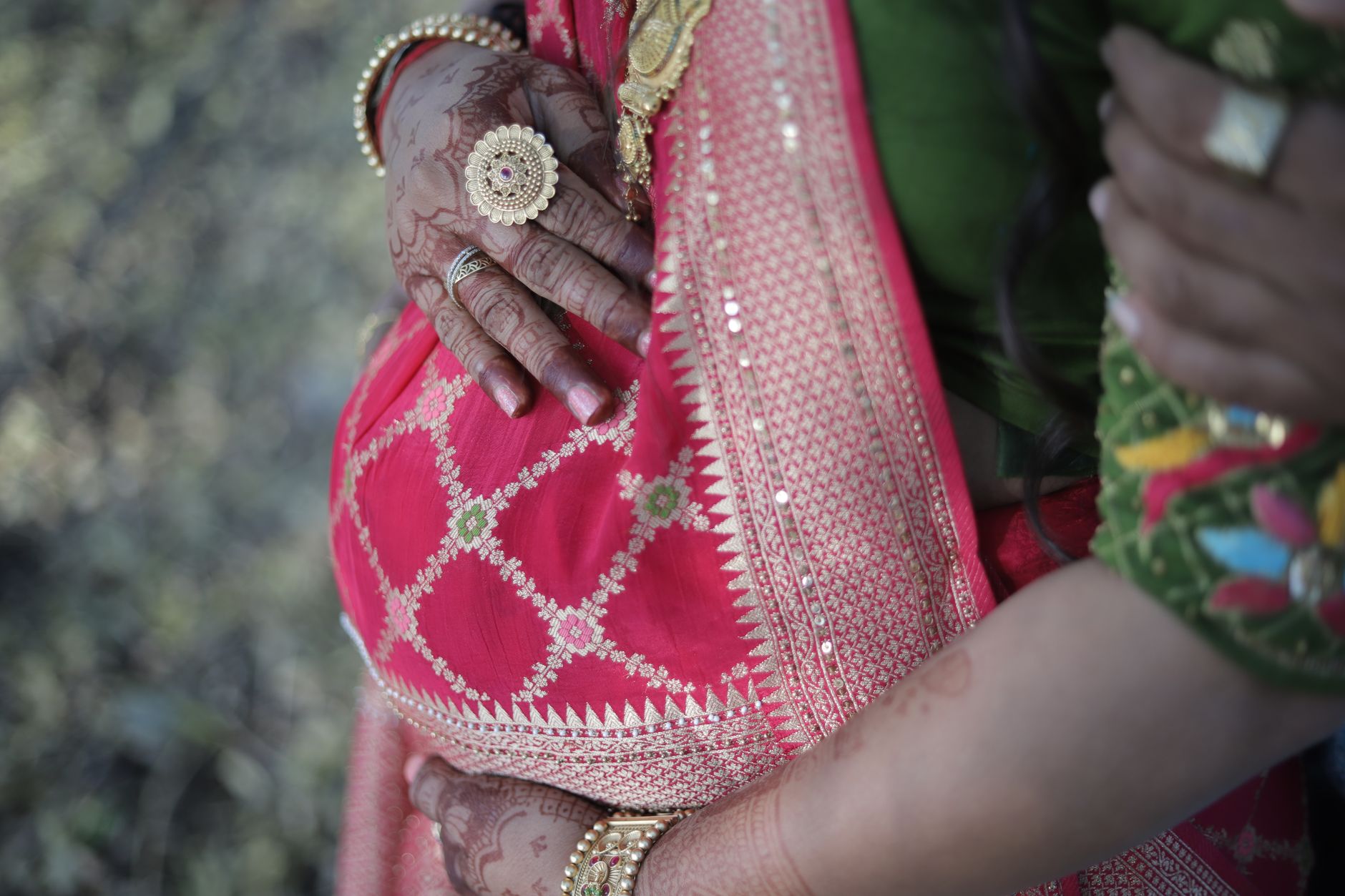 Close-up of a pregnant Indian woman in a pink and gold traditional sari, hands resting on her belly, with a family member's hand nearby