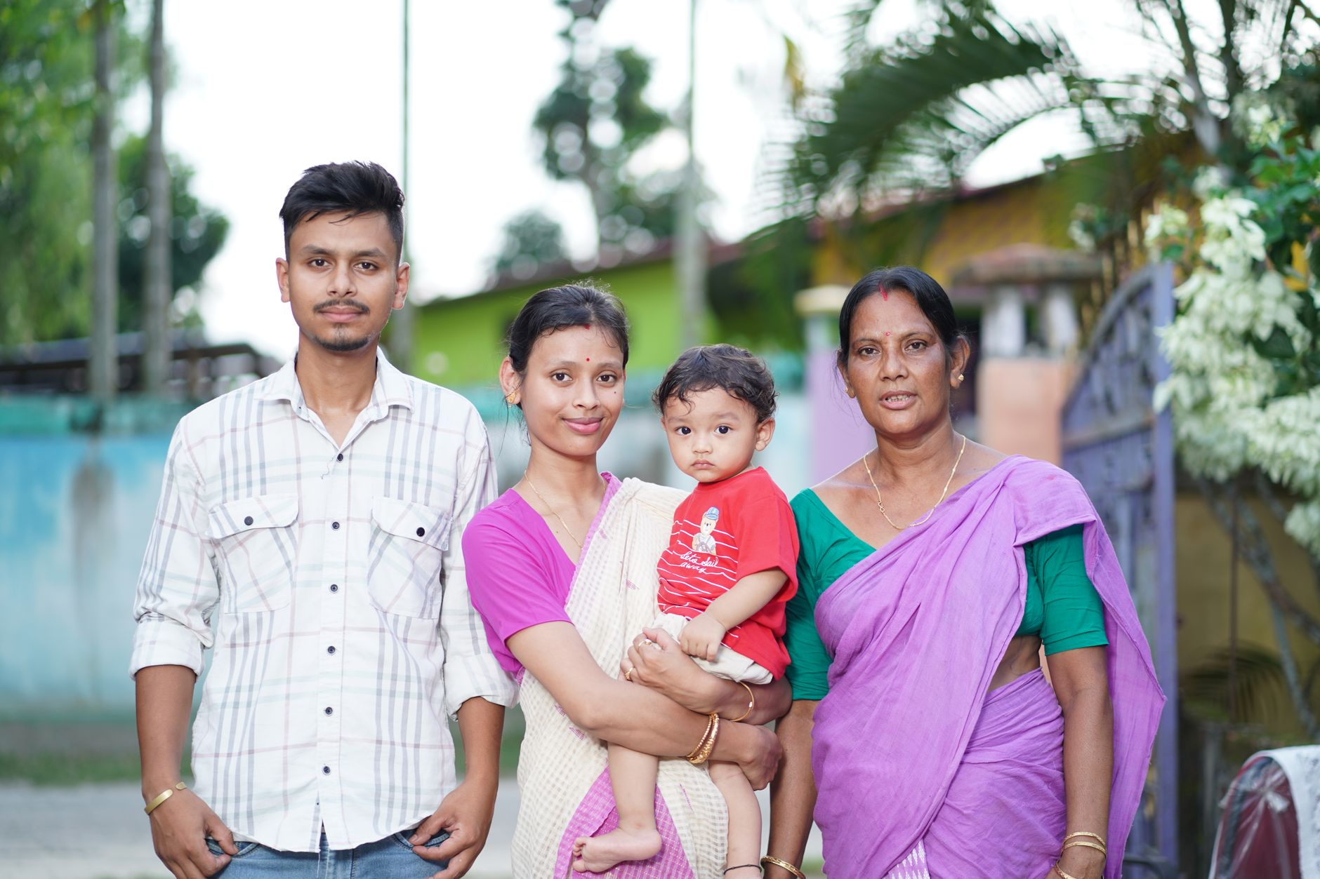 Indian family — young father, mother holding a baby, and grandmother — standing together outside their home in a clean, green neighbourhood
