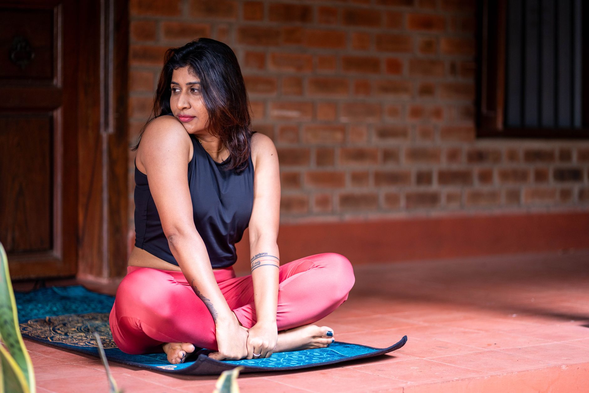 An Indian woman in workout clothes sitting cross-legged on a yoga mat on a home porch, mid-stretch