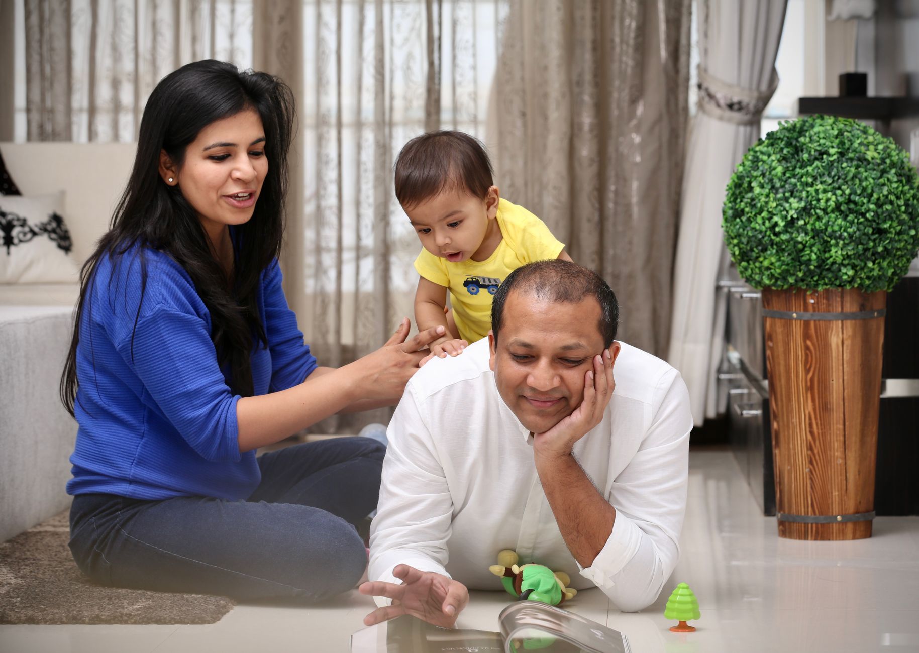 An Indian mother and father playing on the floor with their toddler in a bright, modern living room — no balloons or party decorations, just family time
