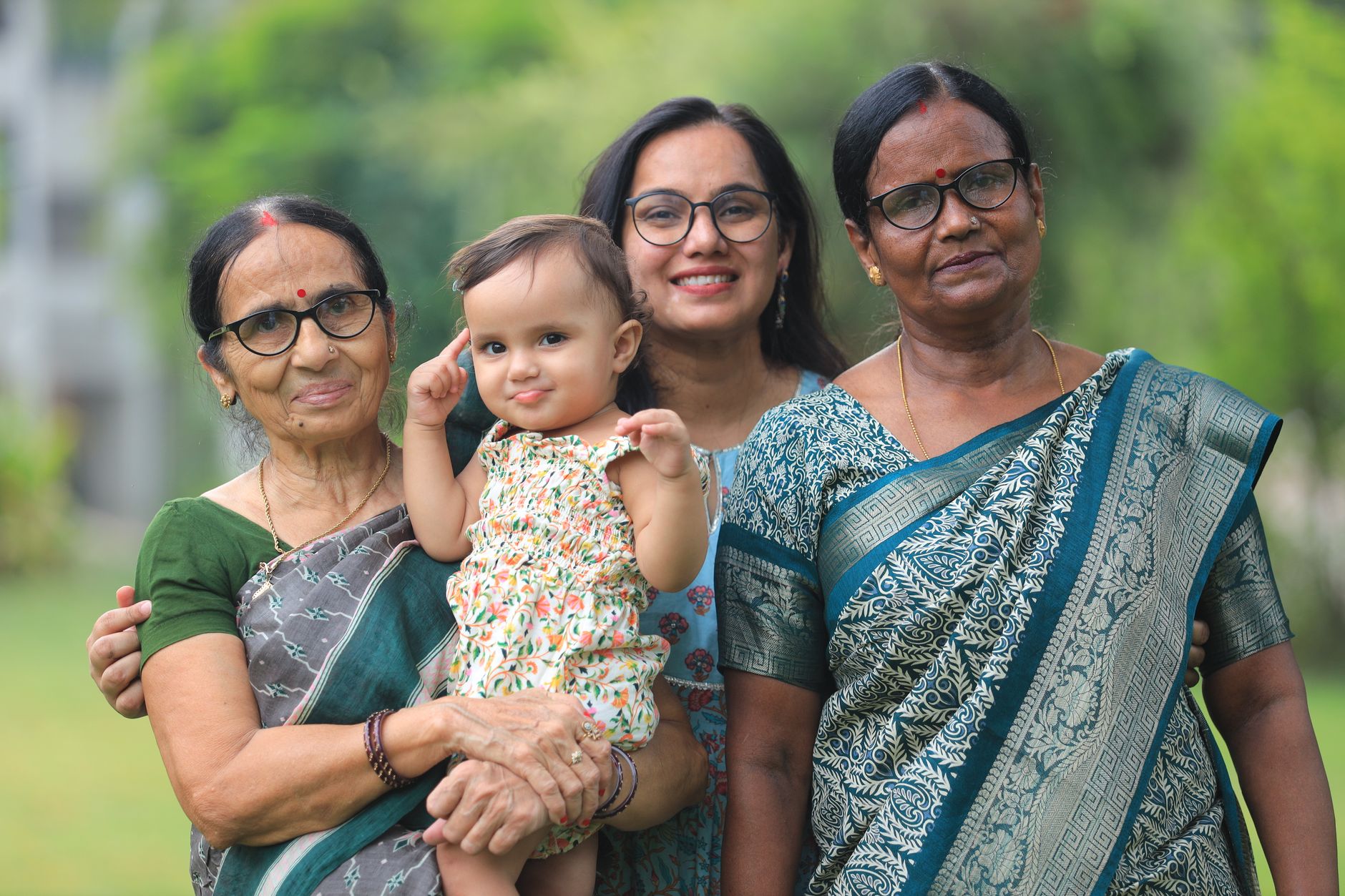An Indian mother smiling and holding her baby, standing with two older women in sarees in a garden setting
