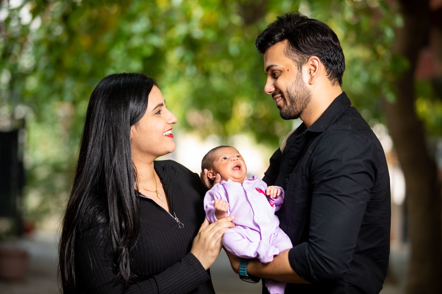 An Indian father and mother stand close together outdoors, both smiling down at their newborn baby held between them