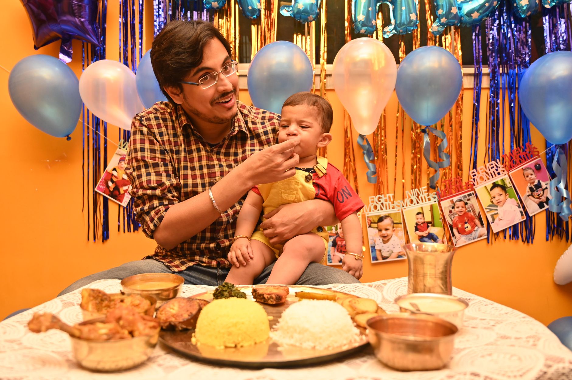 An Indian father feeds his baby son a plate of rice, dal, and chicken curry during a home celebration