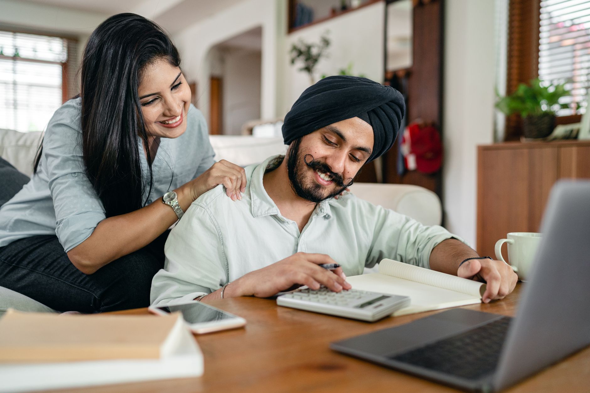An Indian husband and wife going through their insurance paperwork together at home, calculator and notebook on the table, the wife's hand resting supportively on his shoulder