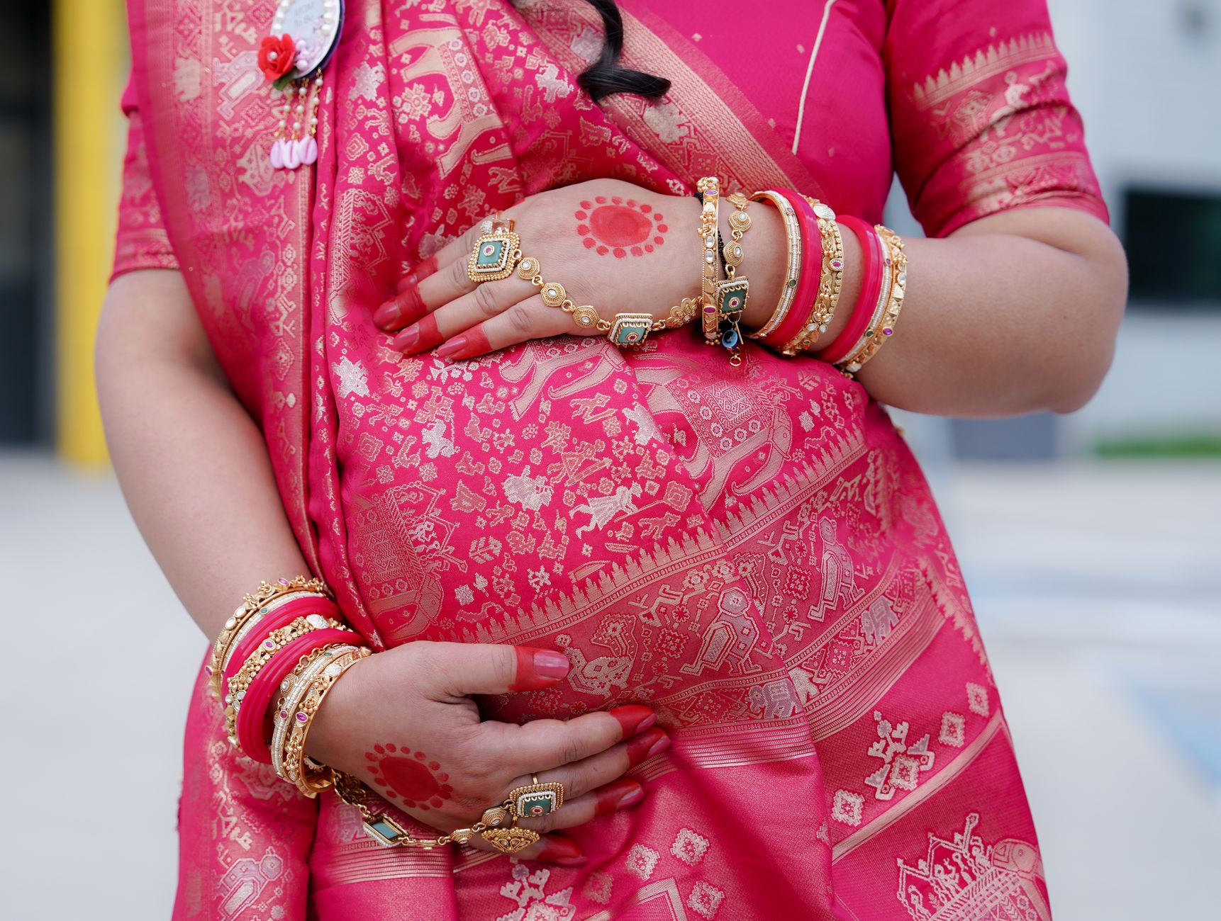 Close-up of a pregnant woman in a pink Indian sari, her hands adorned with bangles, rings, and a small henna design