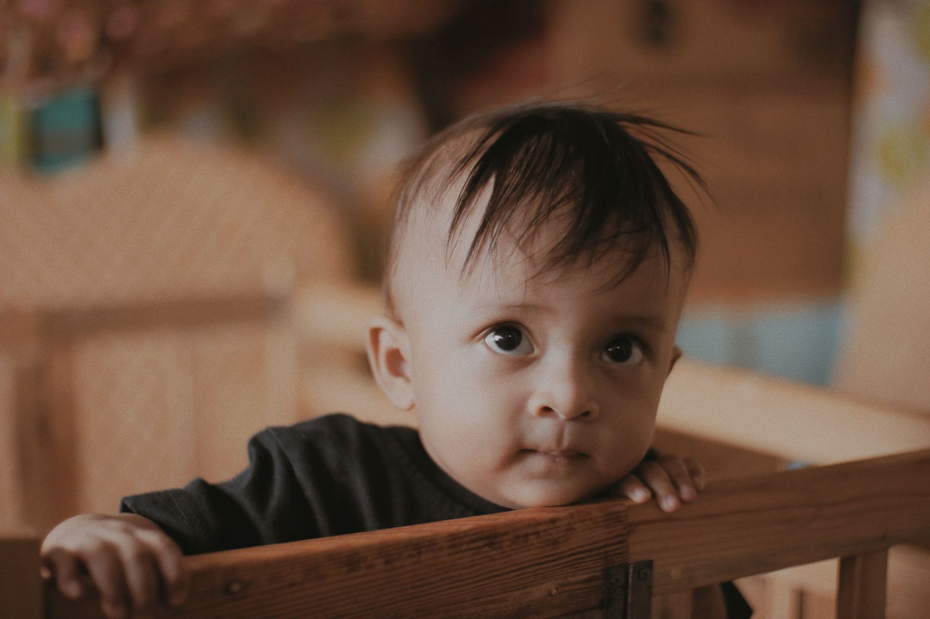 A baby with brown eyes stands gripping the rail of a wooden crib, looking alert rather than sleepy