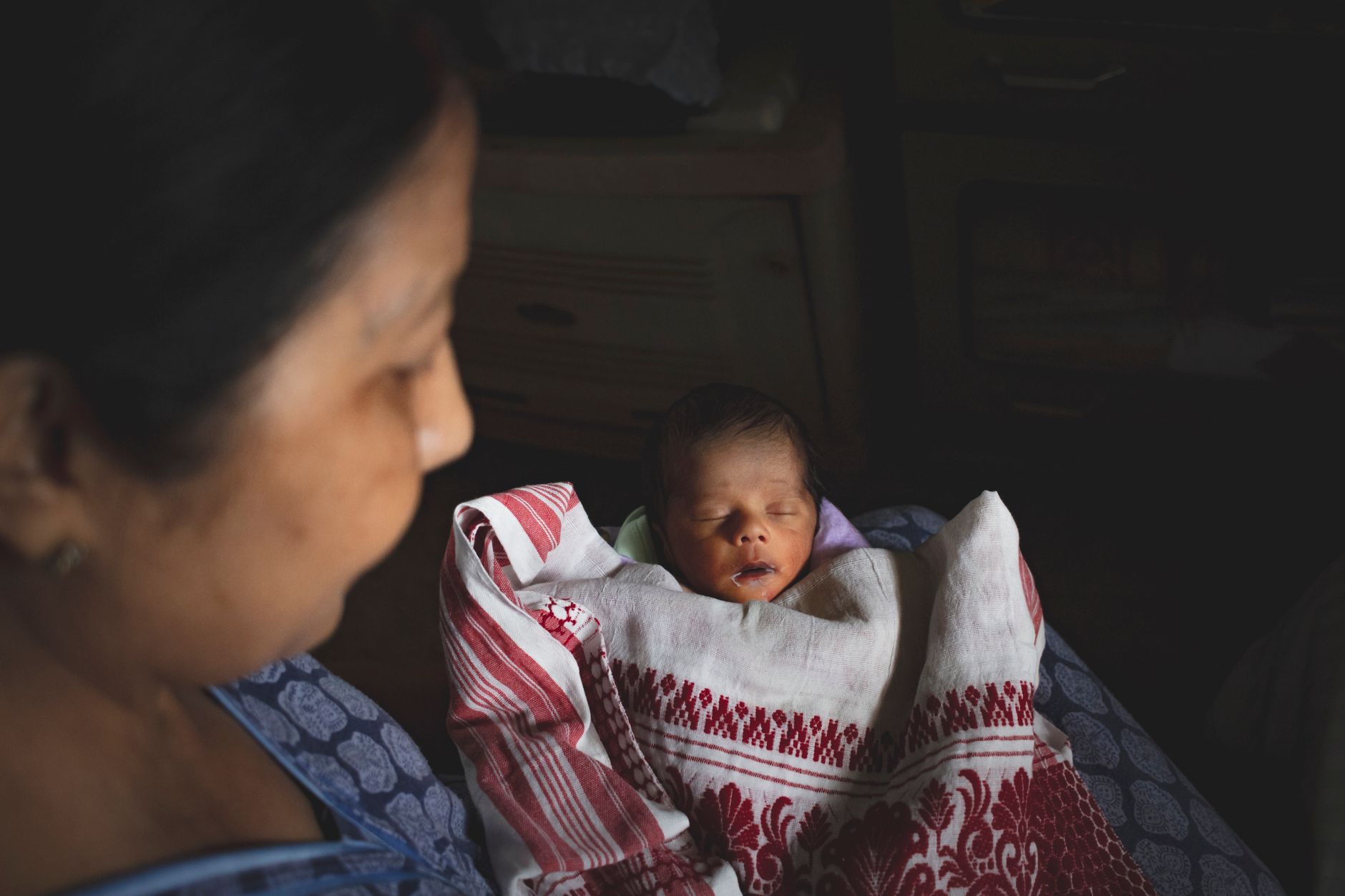 An Indian mother gazing tenderly at her sleeping newborn, who is wrapped in a traditional woven gamusa cloth, in a dimly lit bedroom at night