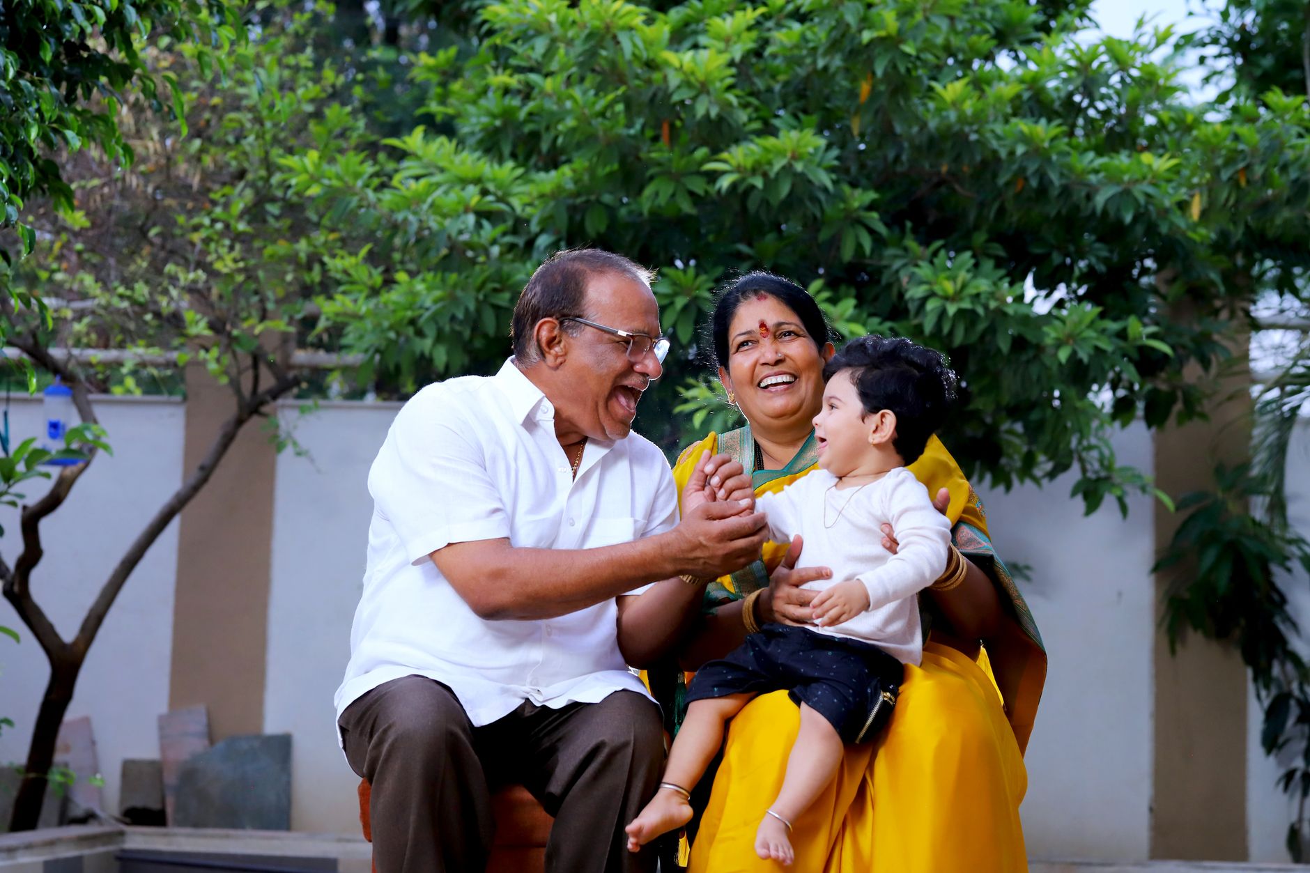 An Indian grandmother holds a laughing toddler while a grandfather leans in smiling, in a green garden