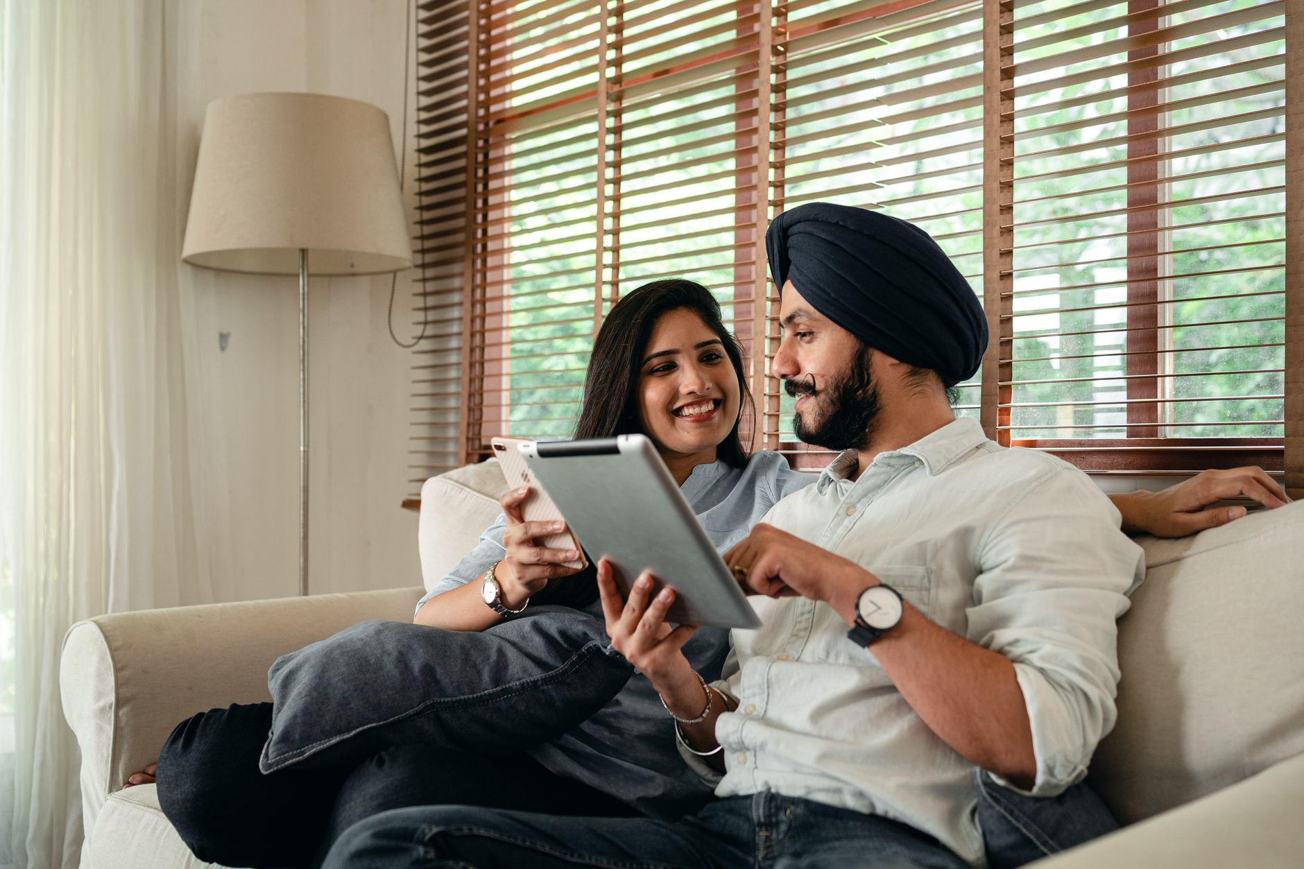 An Indian couple sitting together on a sofa at home, the husband in a turban pointing at a tablet while his wife holds her phone and smiles