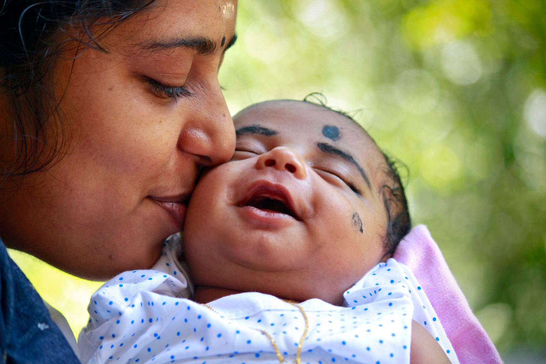 An Indian mother closing her eyes as she kisses her smiling newborn baby, held close outdoors