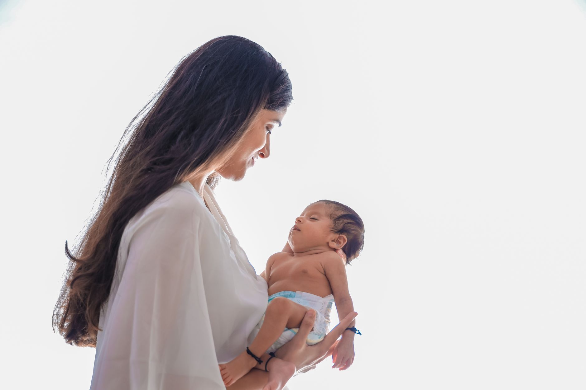 South Asian mother in white flowing clothing holding her newborn baby tenderly with affection in a bright clean setting