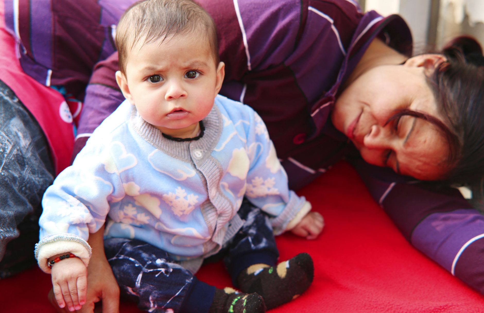 An exhausted Indian mother rests her head down while her alert baby sits beside her on a mat at home