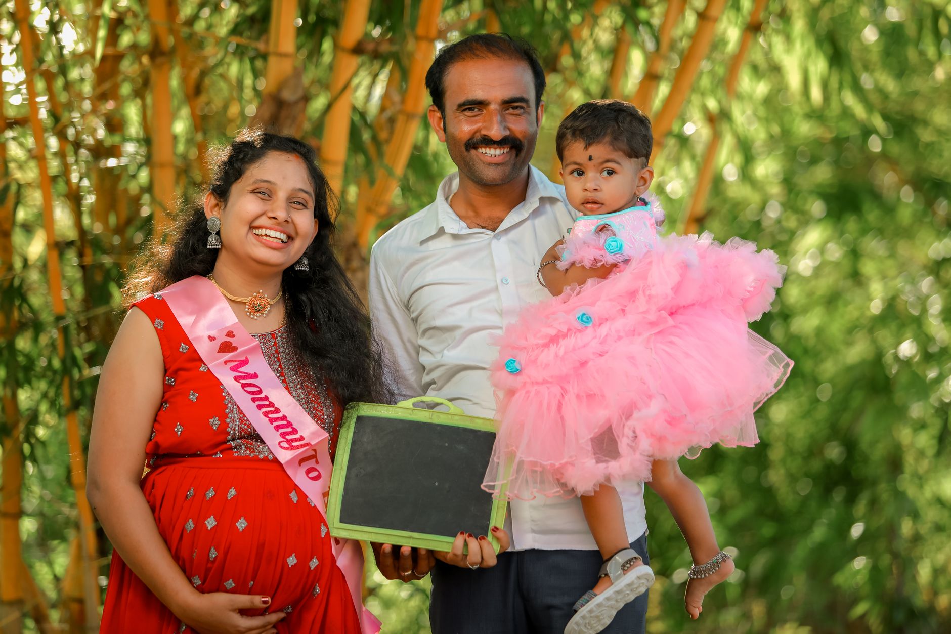 Smiling pregnant Indian mother in a red saree with a 'Mommy to be' sash, standing next to her husband who is holding their toddler daughter outdoors