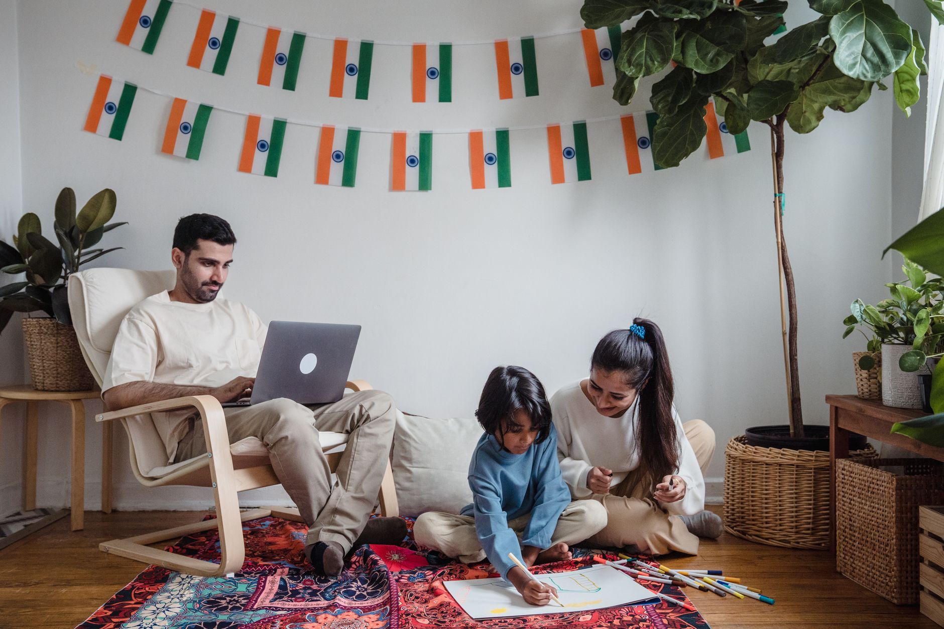 An Indian father works on his laptop while a mother sits on the floor helping her preschool-age daughter draw on paper, in a modern living room decorated with Indian flag bunting.