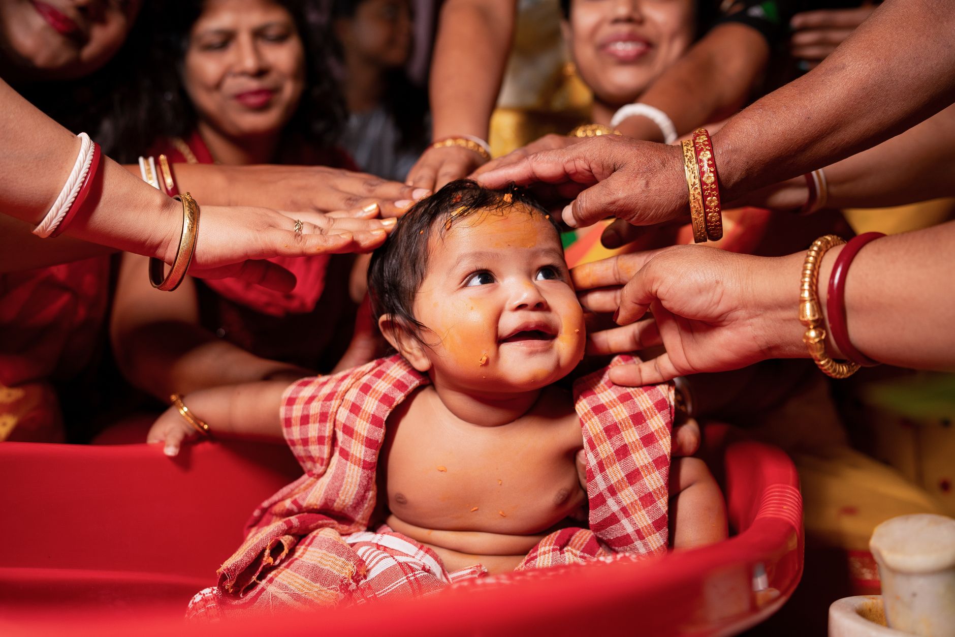 A baby in a red bath tub with turmeric paste on their skin, surrounded by the hands of several relatives blessing them during a traditional Indian family ceremony