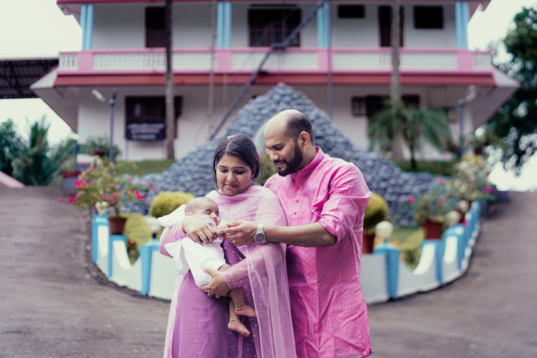An Indian mother in violet salwar kameez and father in pink kurta standing together outdoors, both looking at their newborn baby held in the mother's arms
