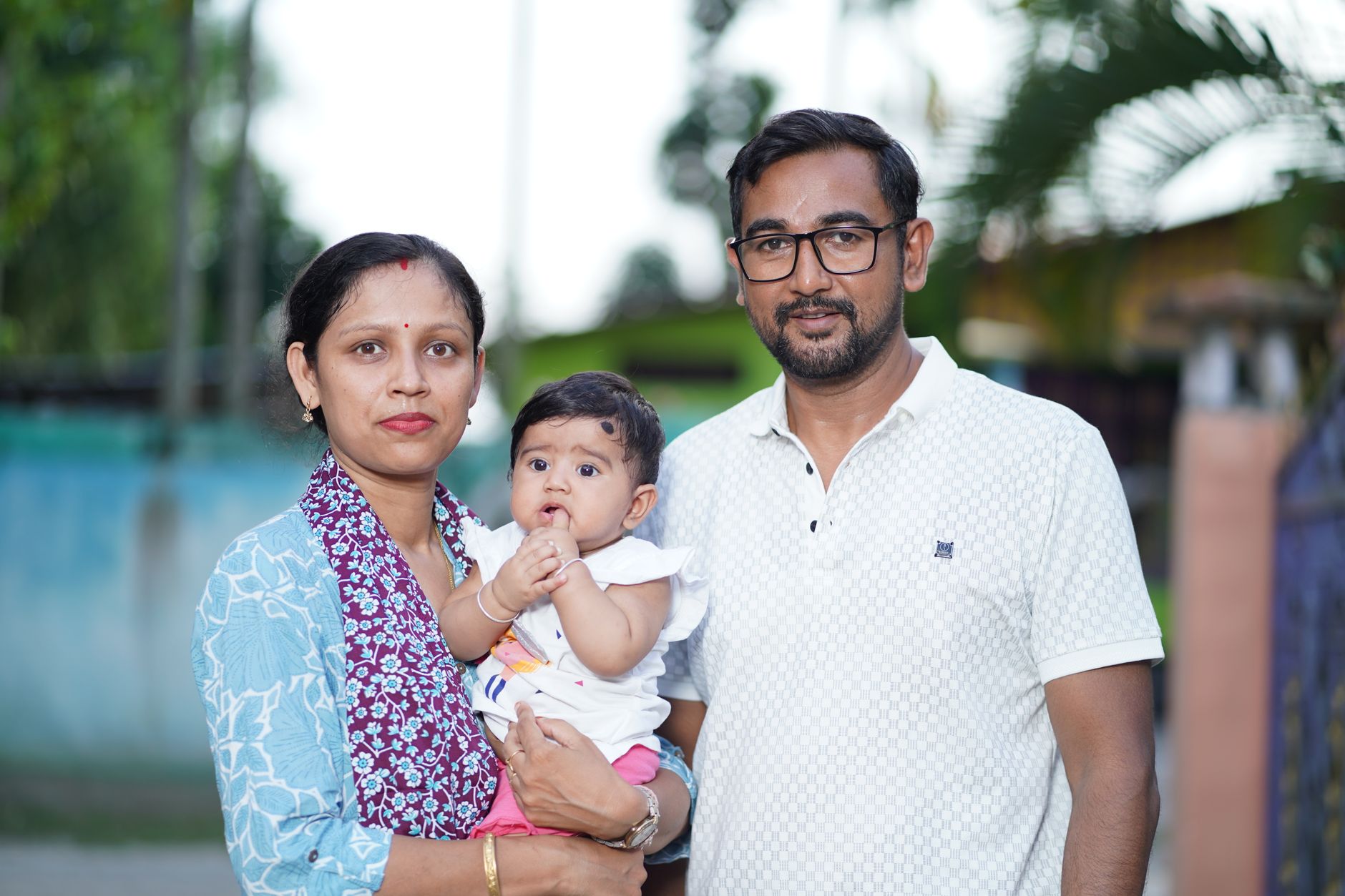 An Indian mother and father standing outdoors together, the mother holding their baby, both parents dressed neatly against a green garden background