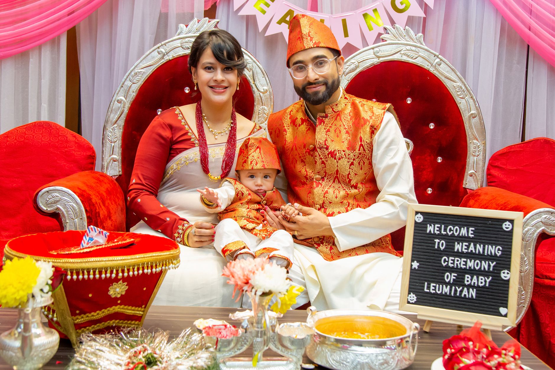 An Indian mother and father, dressed in festive traditional attire, hold their baby at a joyful family weaning ceremony marking the start of solid foods