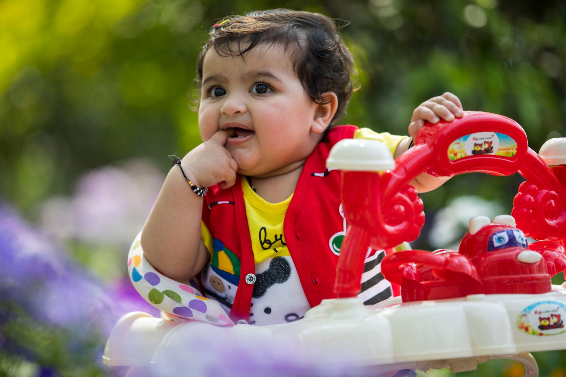 Indian baby around 8–10 months old seated in a colorful stroller outdoors, putting finger in mouth with a bright smile — a classic teething sign — and wearing a traditional black thread bracelet