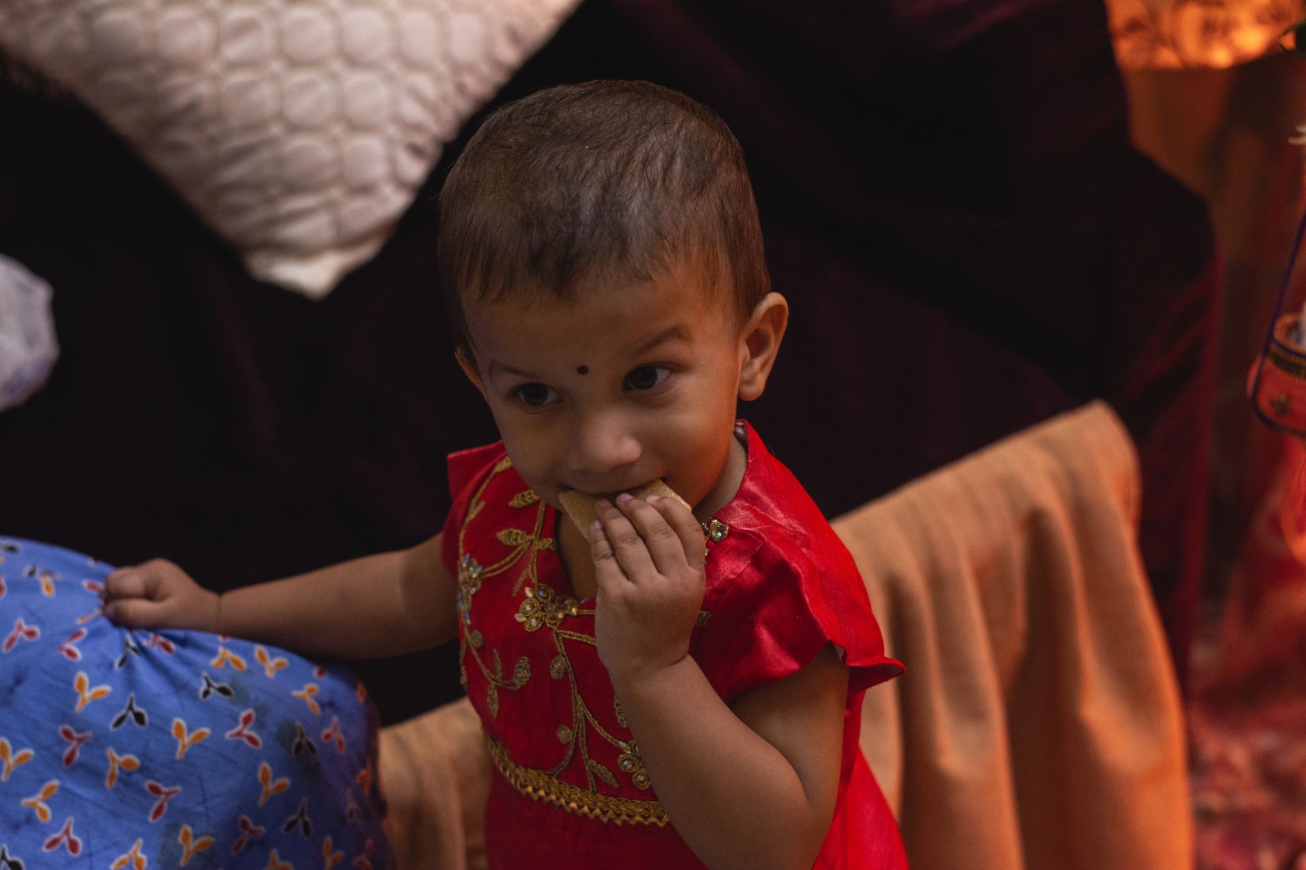 An Indian toddler in a festive red dress eating a snack held up to her mouth