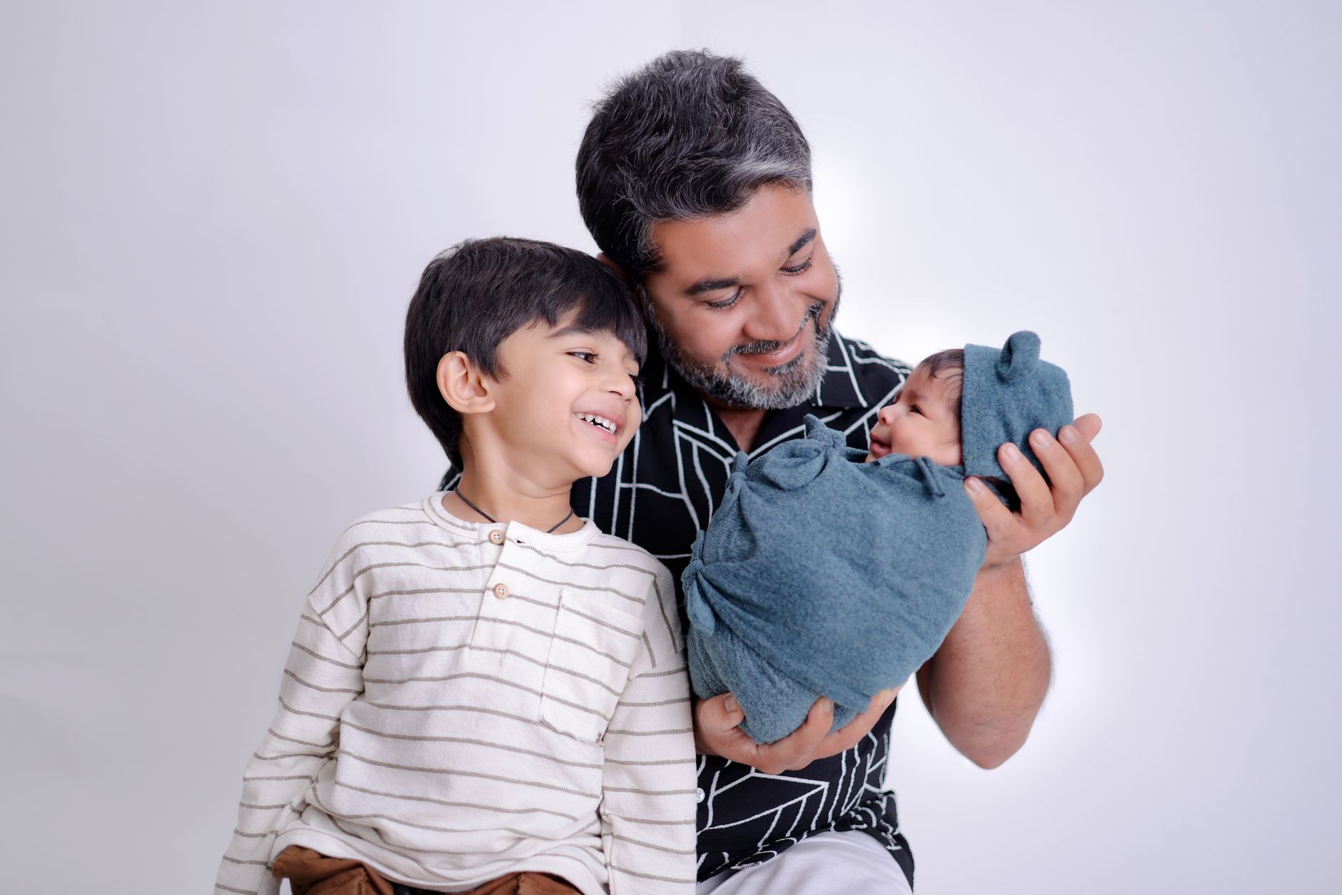 An Indian father smiling down at his newborn, wrapped in a teal cloth, while his toddler son stands beside him laughing, studio portrait