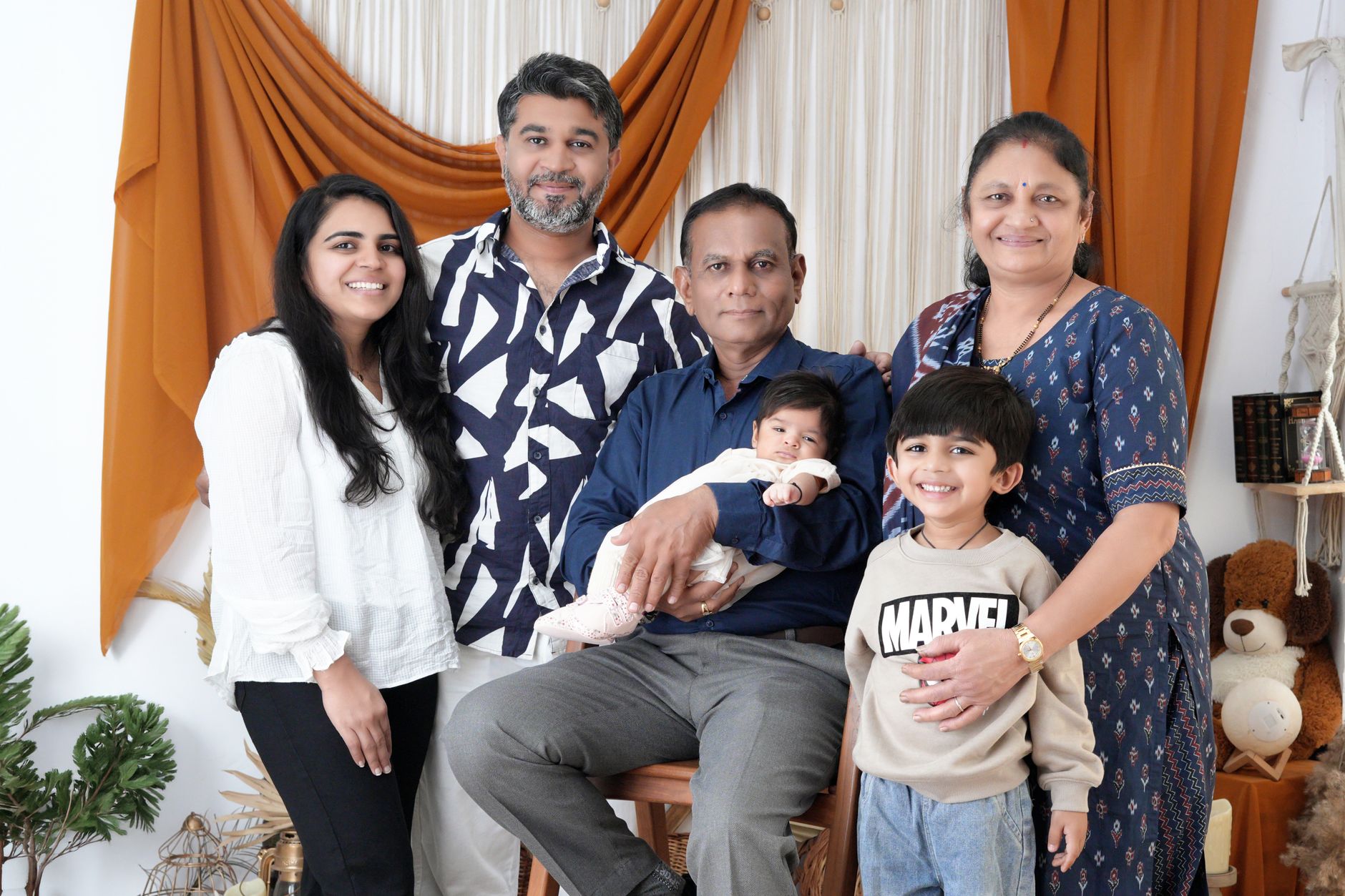 Three generations of an Indian family — grandparents, parents, and children including a baby — sitting together indoors for a family visit